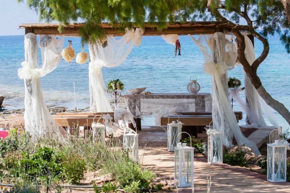 A pathway lined with lanterns leads towards a wooden gazebo decorated with lace against a backdrop of the beautiful sea