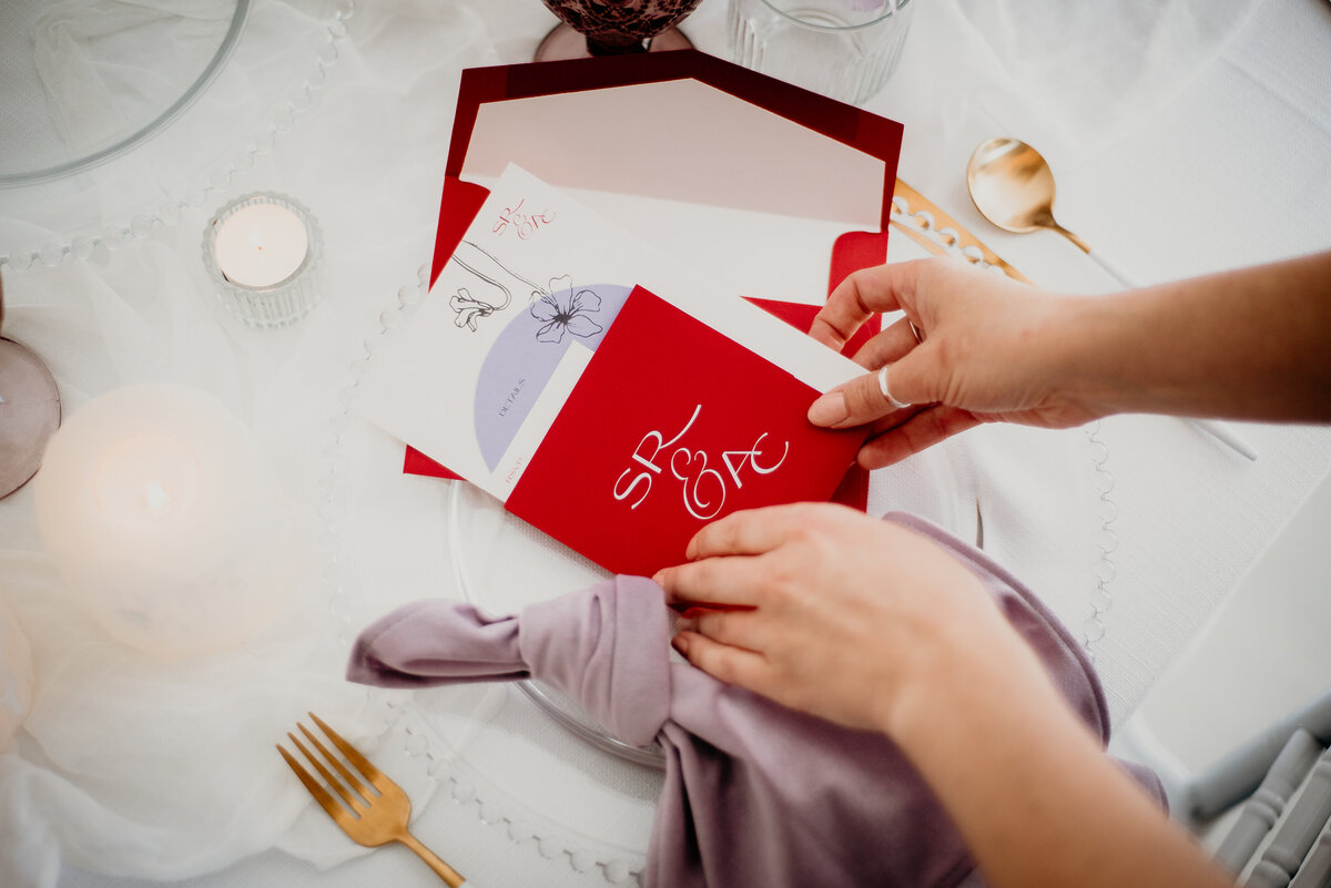 Two hands holding a bright red and purple wedding invitation suite with a paper sleeve