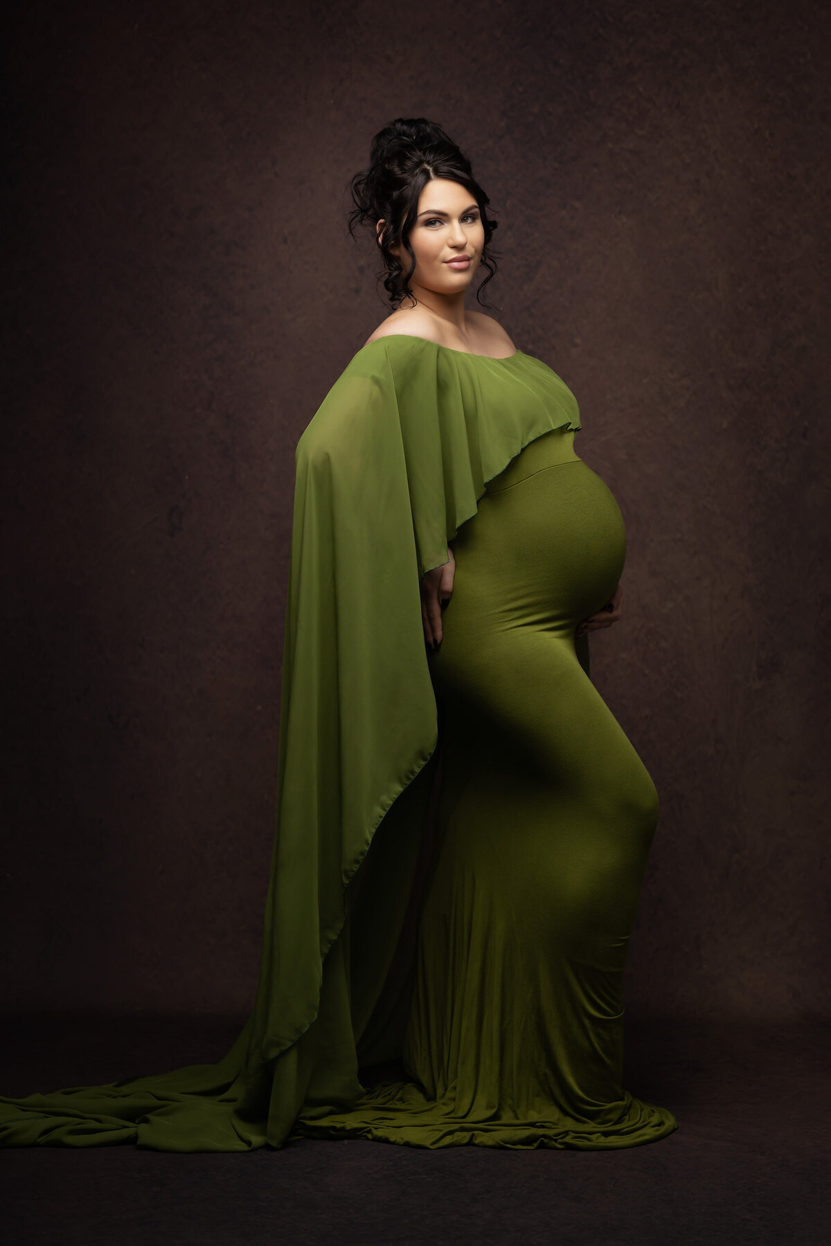 Pregnant woman in a flowing green gown, softly lit against a rich, textured backdrop.