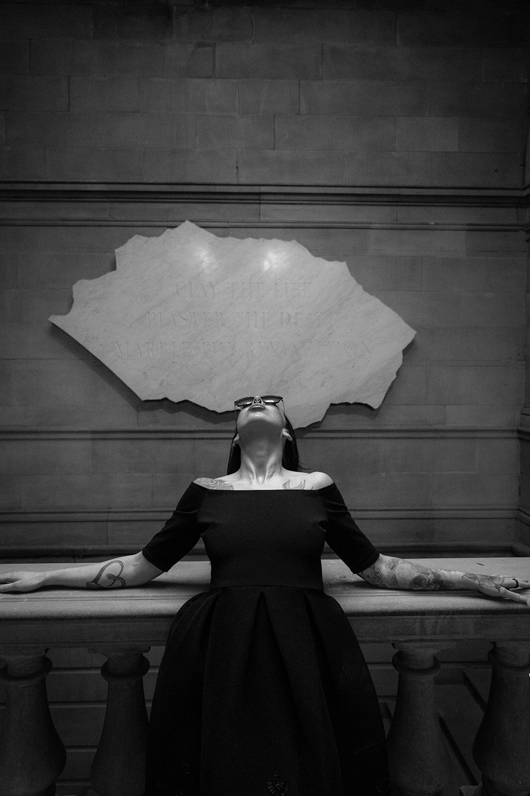 A lady in a black dress and sunglasses leans back against museum steps and lets the sun light up her face