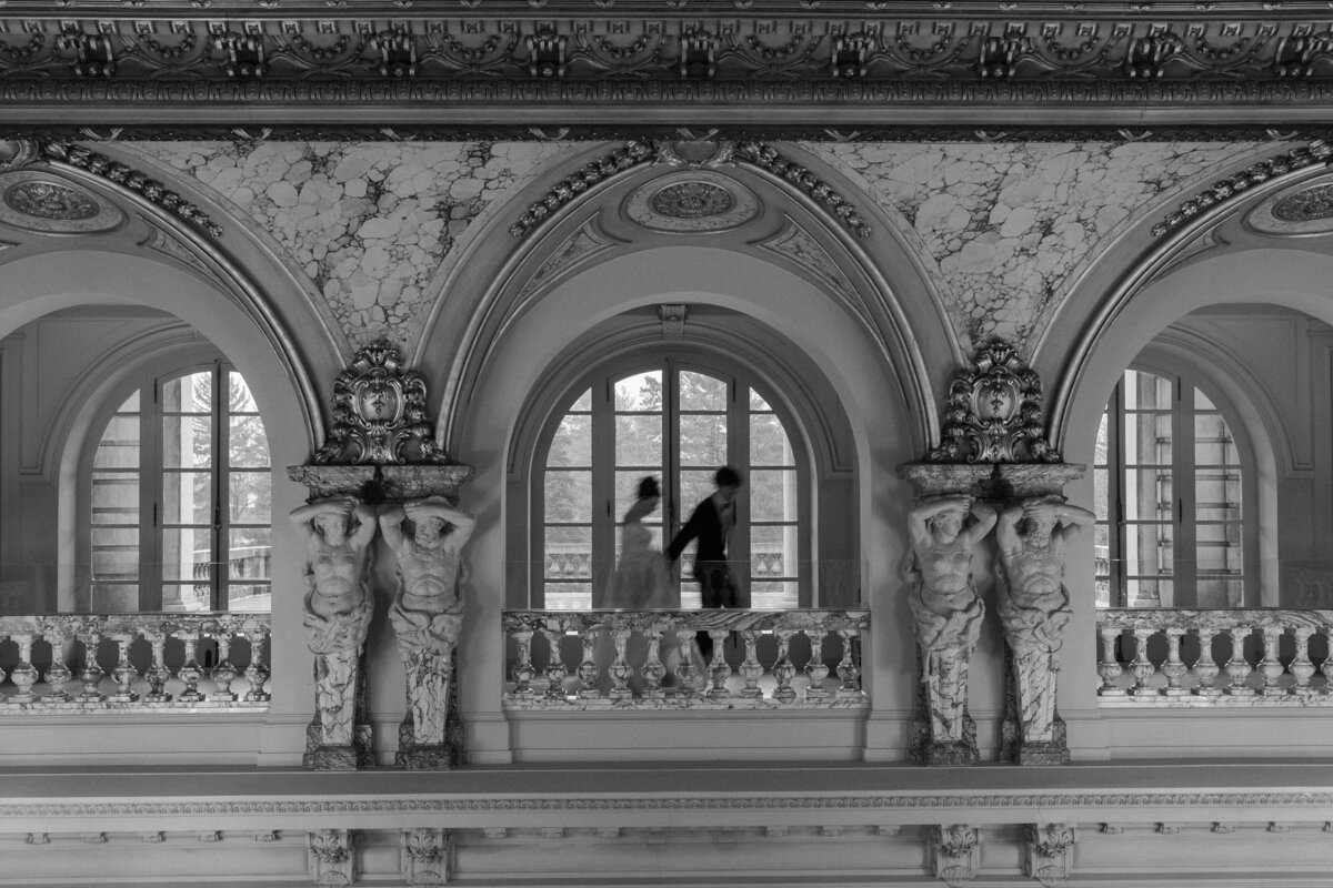 A black-and-white image of a couple walking hand-in-hand along a balcony, flanked by ornate columns and arched windows.