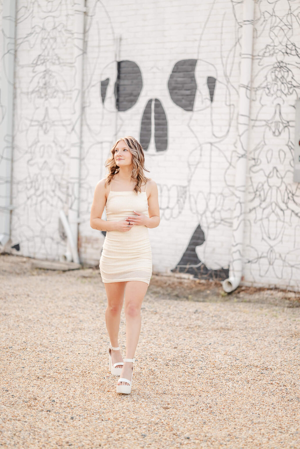 A high school senior, wearing an off-white body con dress and shoes, walks away from a skull mural. She is having her senior session with Chesapeake Senior Photographer, Justine Renee Photography.
