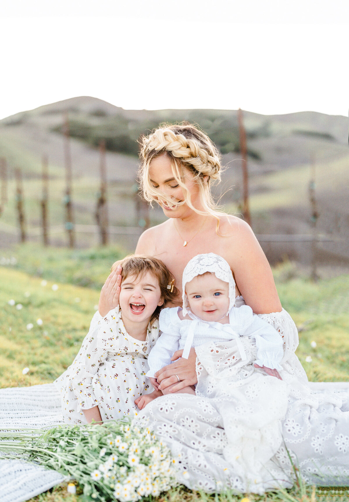 A mother in a white gown with braided hair crown sits in an open field with her toddler daughter wearing a floral white dress and baby wearing a white bonnet and gown photographed by Bay Area Family Photographer, Light Livin Photography.