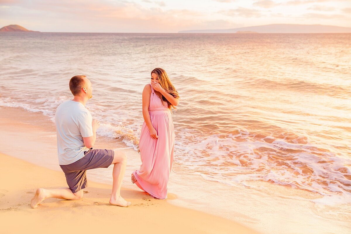 Woman in a pink dress covers her mouth in surprise as her partner gets down on one knee in front of their Maui couples photographer