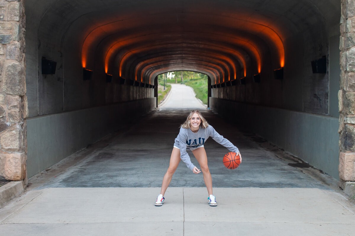 A person wearing a gray sweatshirt and shorts is playing basketball in a tunnel. They are dribbling a basketball while smiling. The tunnel is lit with orange lights on the ceiling, and theres a path visible at the end of the tunnel.