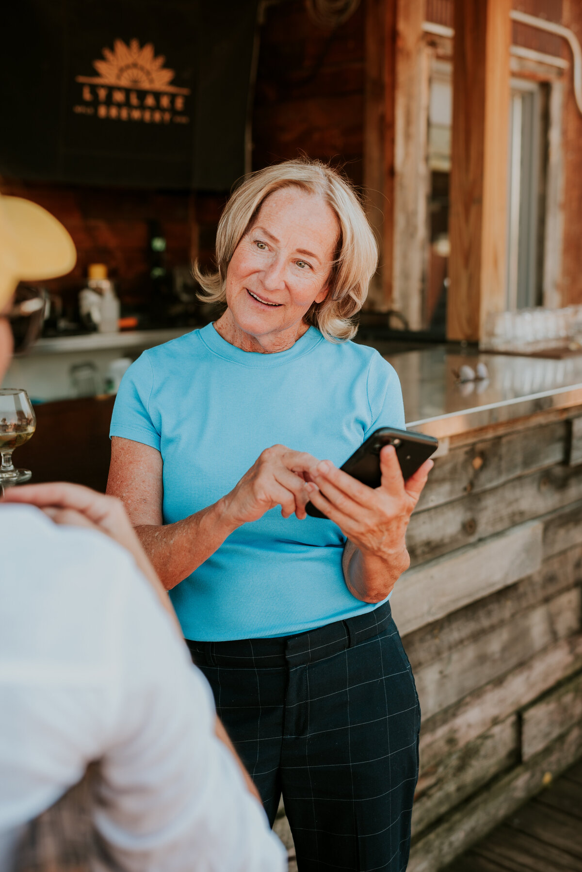 Blonde woman laughing while sitting at a rustic table in a turquoise shirt, perfect for an authentic dating profile.