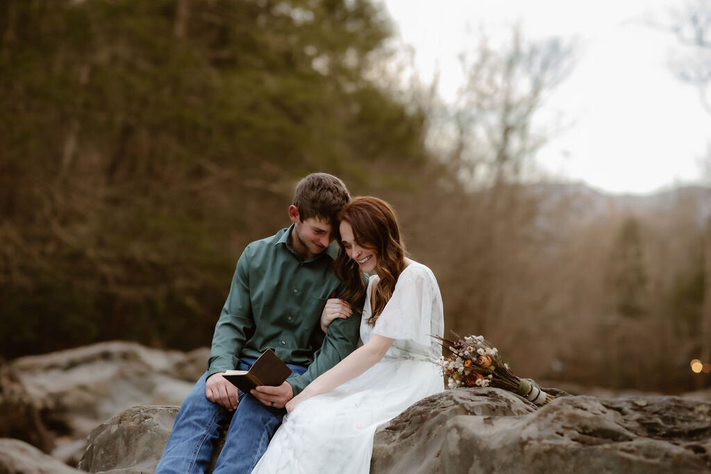 Bride and groom sitting closely together on a large rock at Greenbrier, smiling and reading vows from a small notebook, with a rustic bouquet beside them during their eloping to Gatlinburg ceremony.
