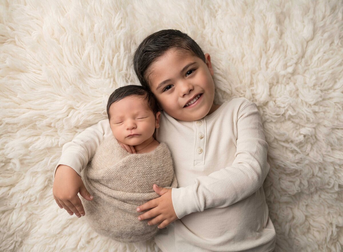 Aerial image. Big brother is captured holding his little brother at his Lake Elsinore newborn photoshoot. Big brother is smiling at the camera while the sleeping newborn is resting in his big brother's shoulder. Captured by best Lake Elsinore newborn photographer Bonny Lynn Photography