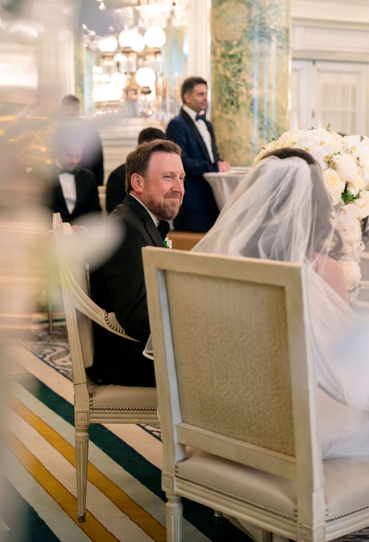 A wedding ceremony in an elegant venue. The groom, wearing a black suit, sits on a chair adorned with a floral arrangement. The bride is next to him in a white veil. Guests in formal attire are in the background.