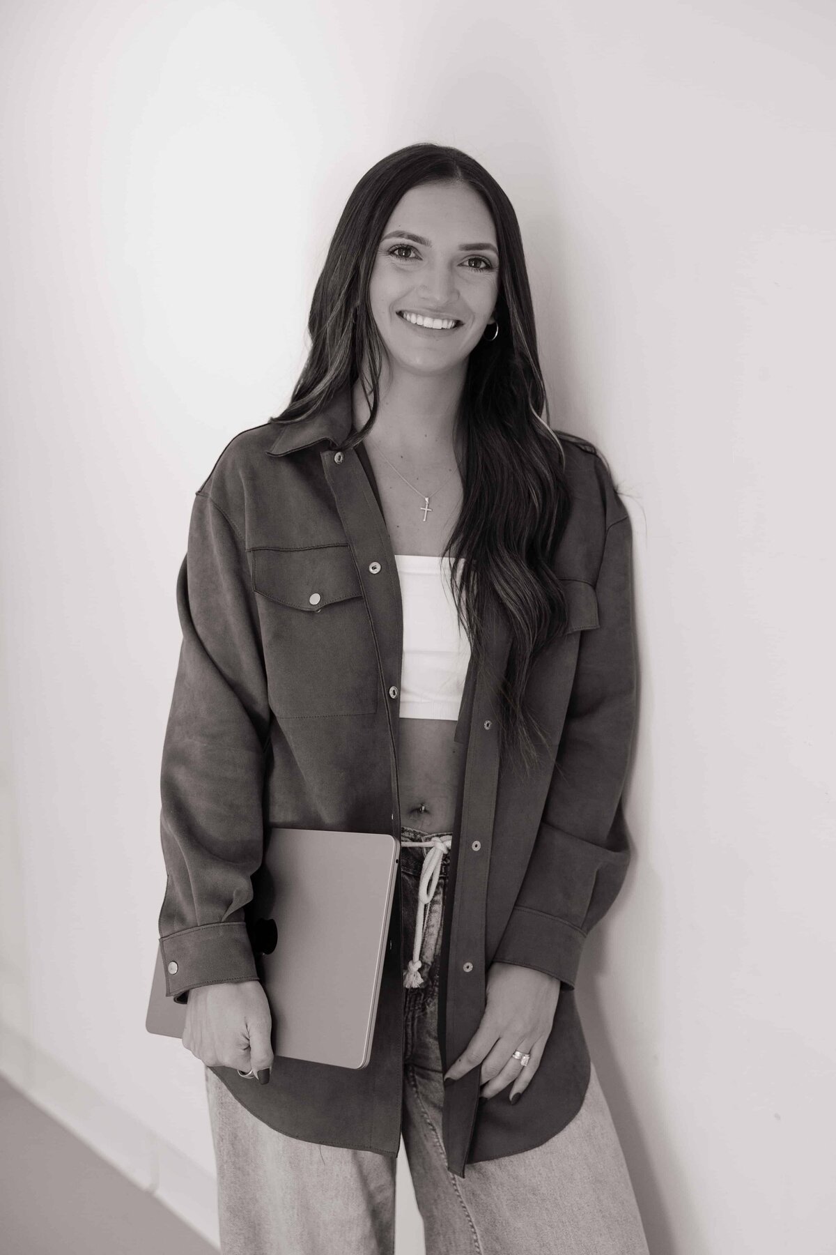 A fitness coach smiles for the camera wearing casual clothing and holding her laptop.