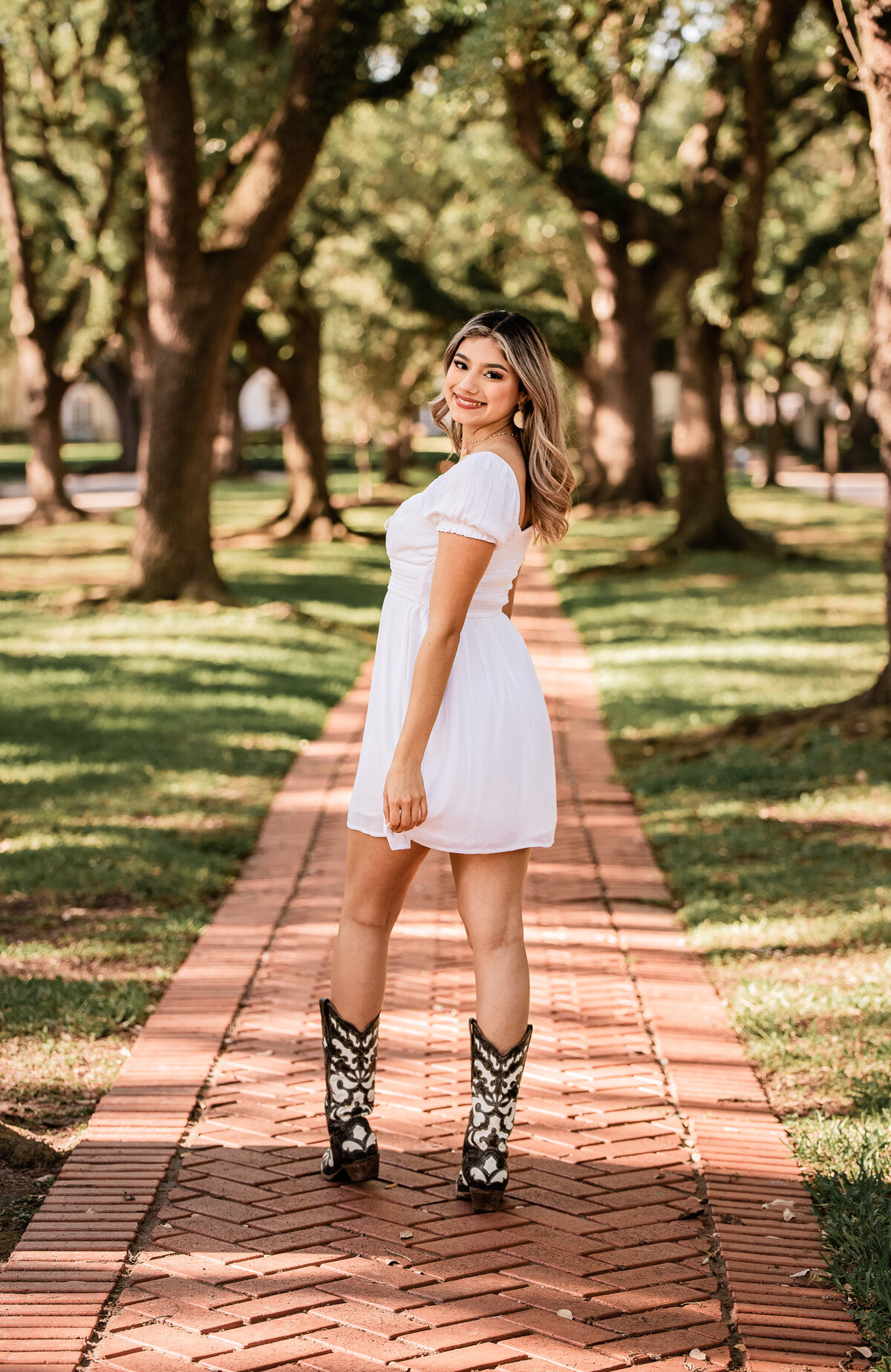 A teen girl wearing a white dress and cowboy boots looks over her shoulder on a red brick walkway.