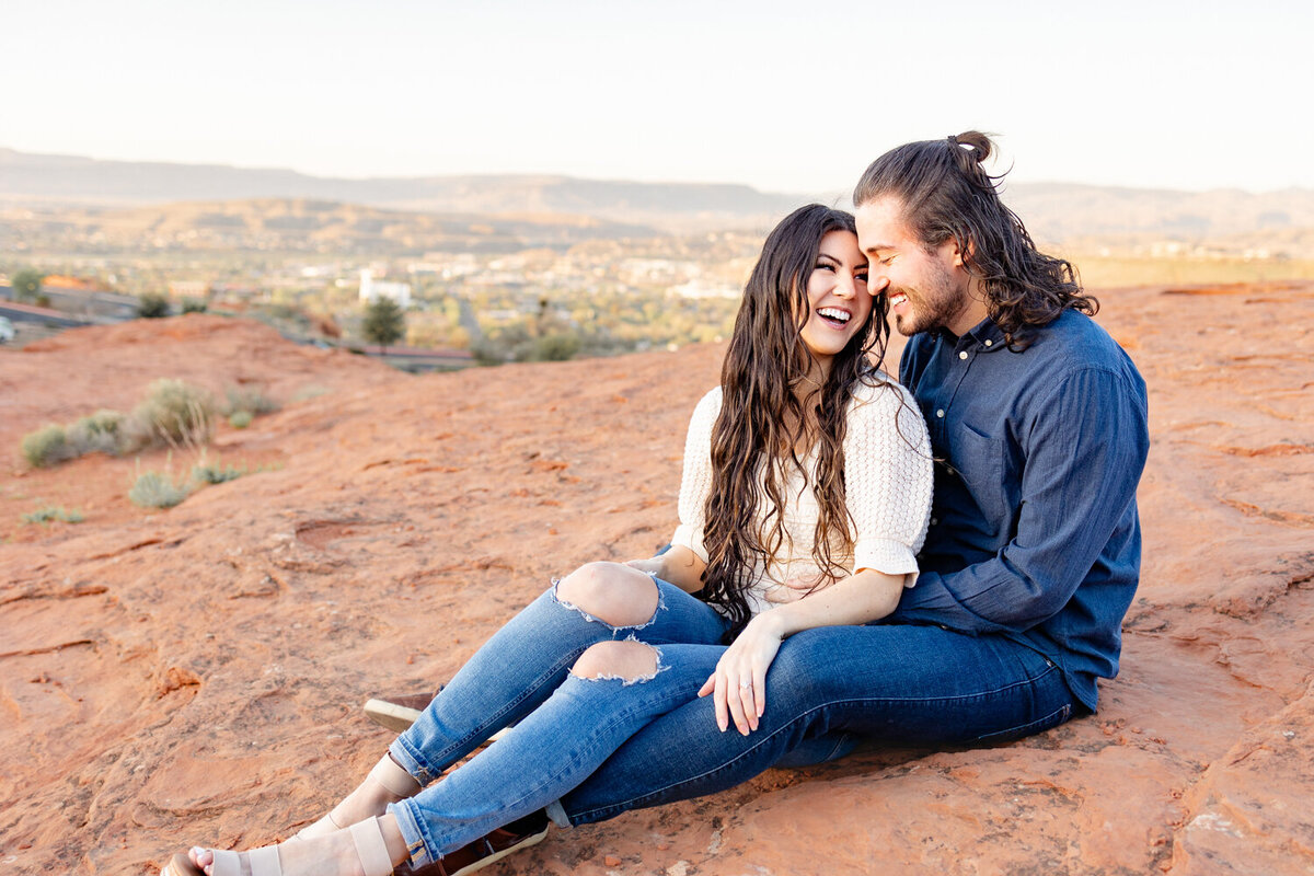 Engaged couple laughing with one another as they snuggle together on the red rocks in St George UT