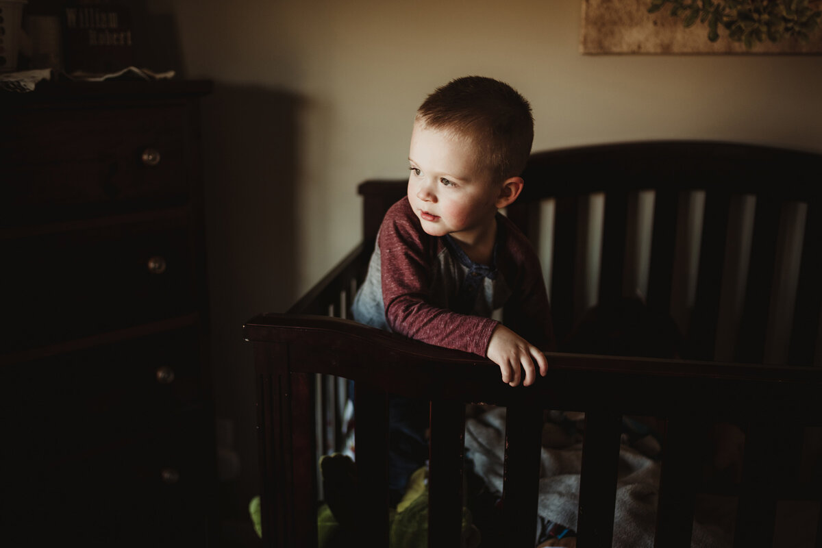 A little boy is standing in a crib while looking out the window in moody light.