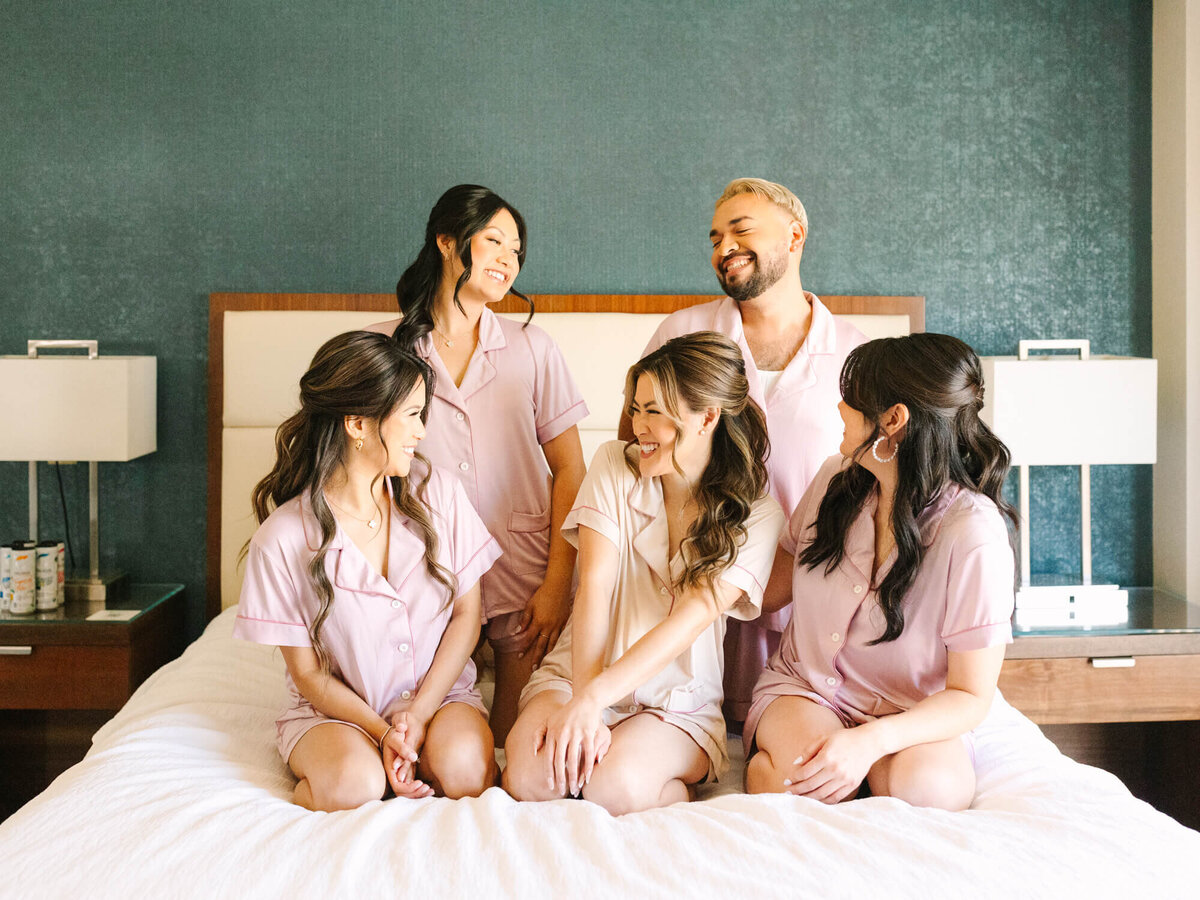 Five people in matching pink pajamas sit and kneel on a bed, smiling and laughing together. The room has teal walls and modern decor, creating a joyful and relaxed atmosphere.