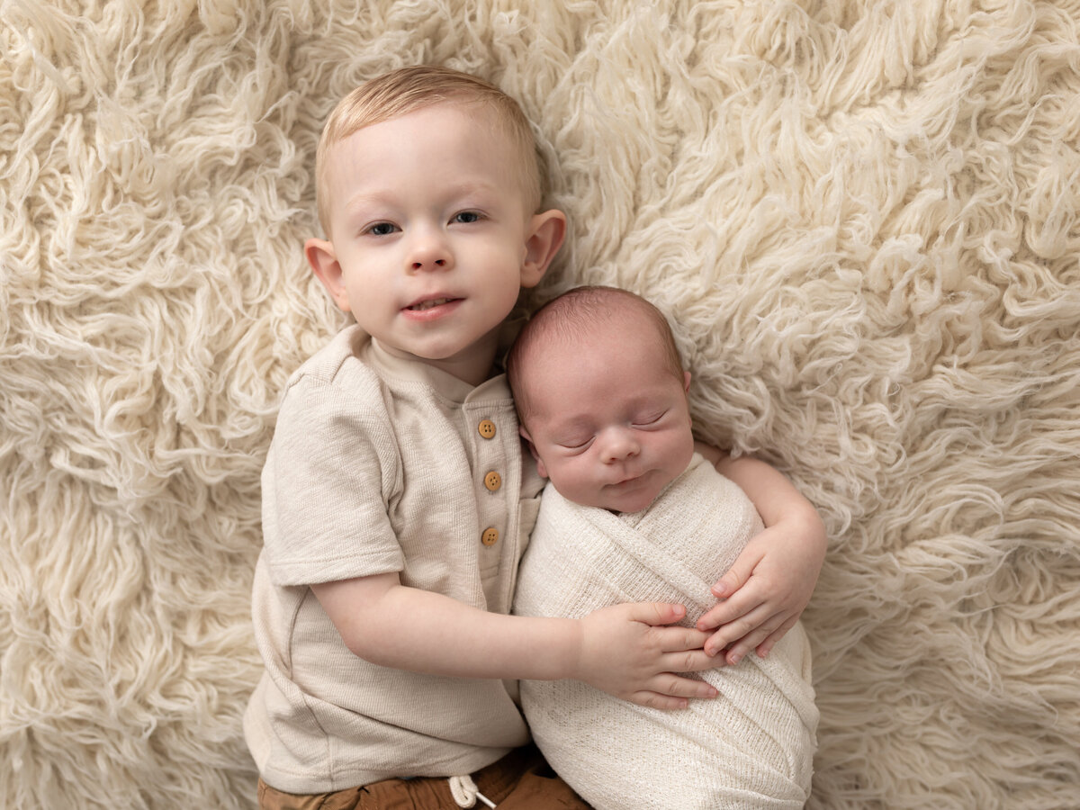little boy in cream shirt holding newborn baby boy in white swaddle