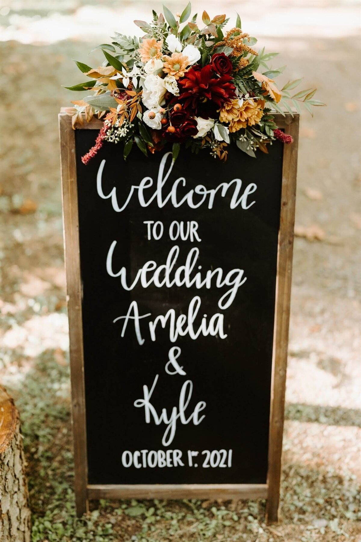 Wedding welcome sign with floral arrangement sitting atop it to welcome guests.