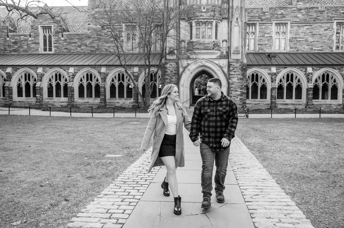 A man and woman walk hand in hand on a path in front of a historic building with arched windows.