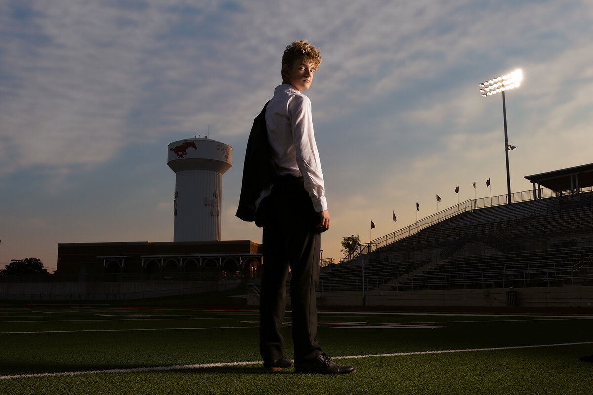 A senior guy stands with his back to the camera, holding his suit jacket over his shoulder in front of Grapevine High School's football stadium. The iconic Mustang water tower and stadium lights create a striking backdrop for this dramatic Dallas senior photography shot.