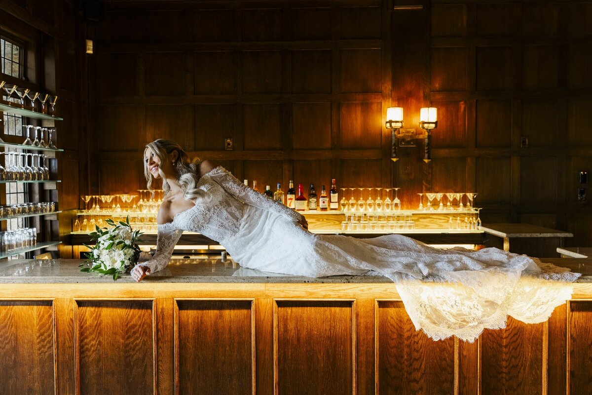 Bride lying across a bar with her gown spilling over the counter in a warmly lit room