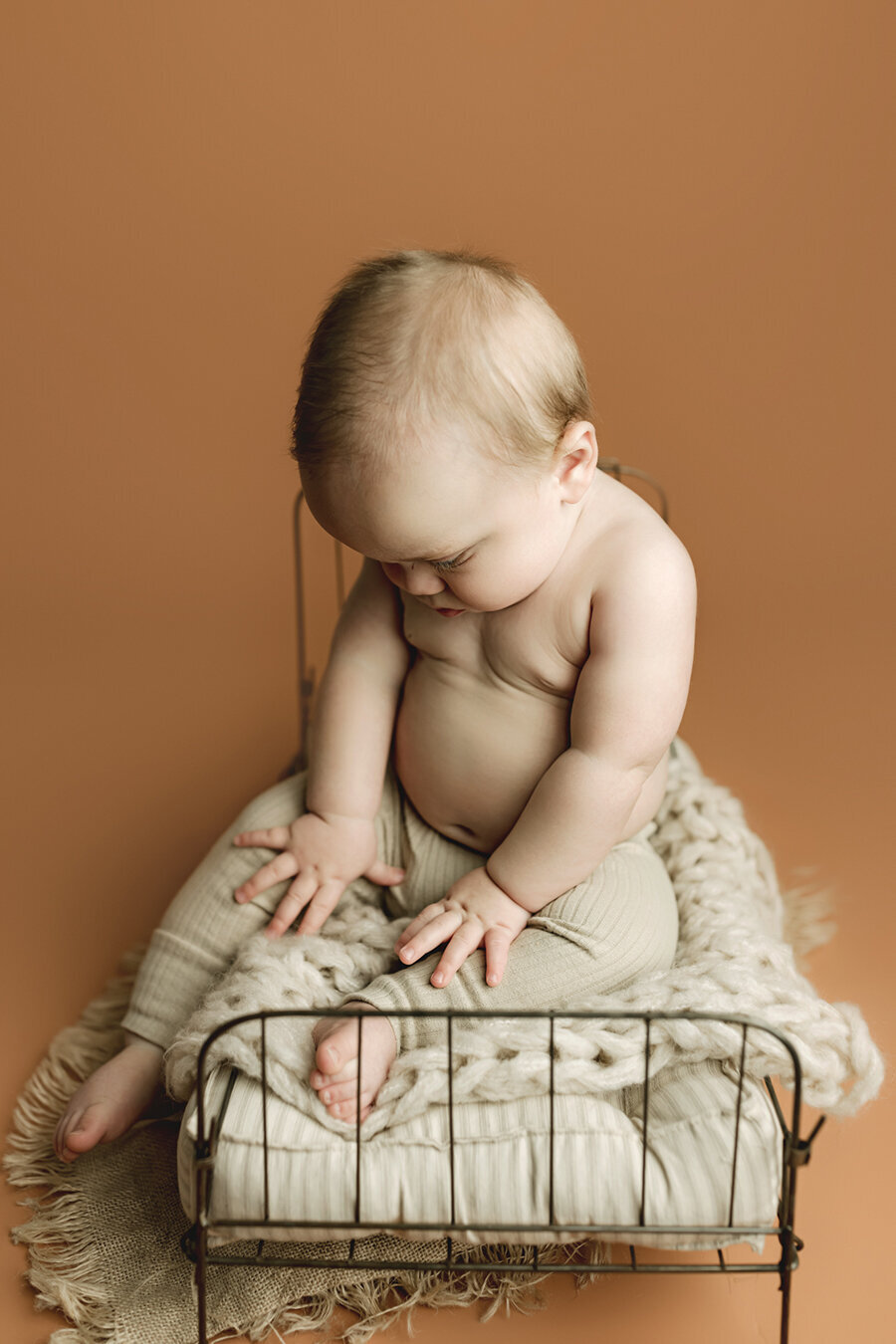 Six month old baby boy looking at his hands while he sits on a little bed with his shirt off