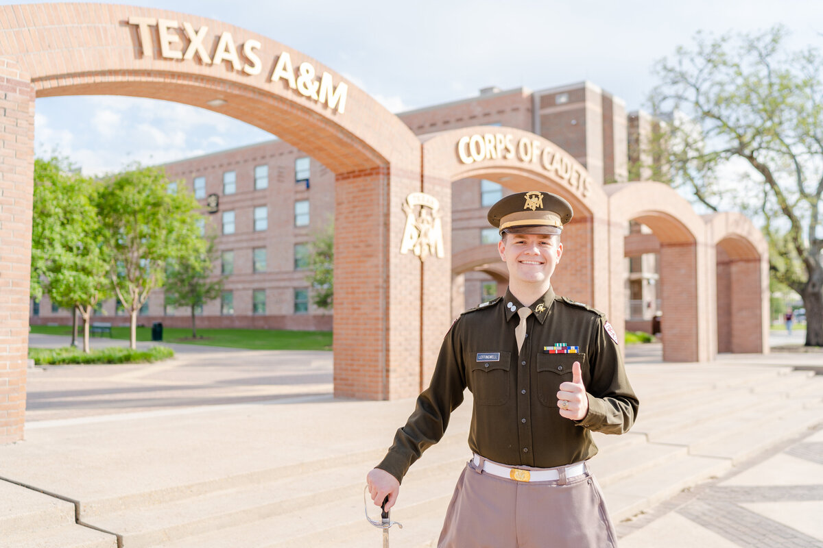Texas A&M senior guy wearing CORPS uniform standing in front of Corps arches and thumbs up gig em