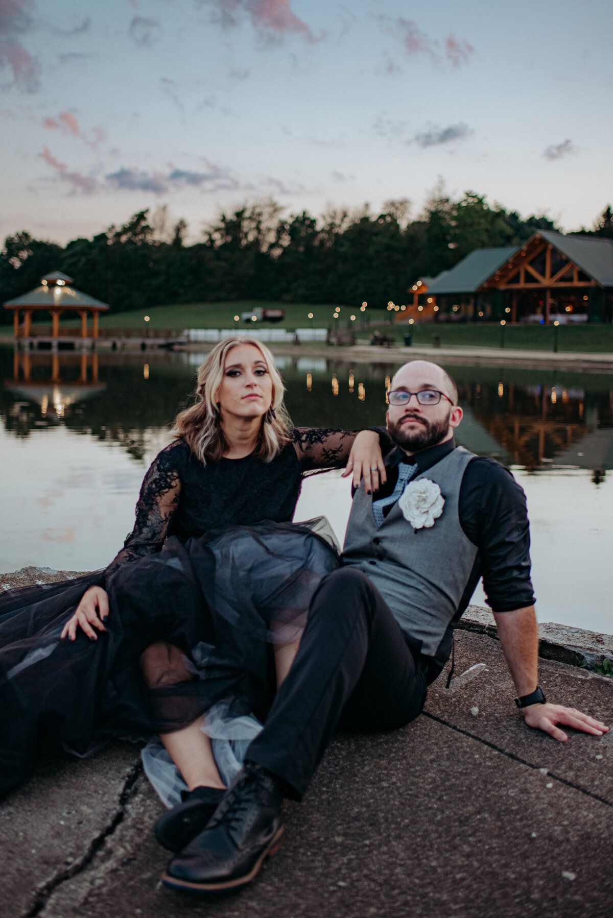 An editorial style photo with alt vibes as the bride in a black dress sits with the groom on the dock of a lake during blue hour portraits.