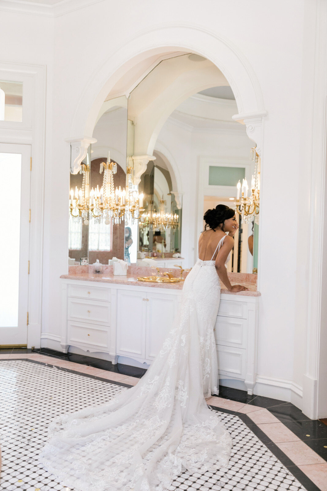 Portrait of bride Nadia looking at herself in the mirror, showcasing the back of her Allure Bridals gown in a beautiful white bathroom