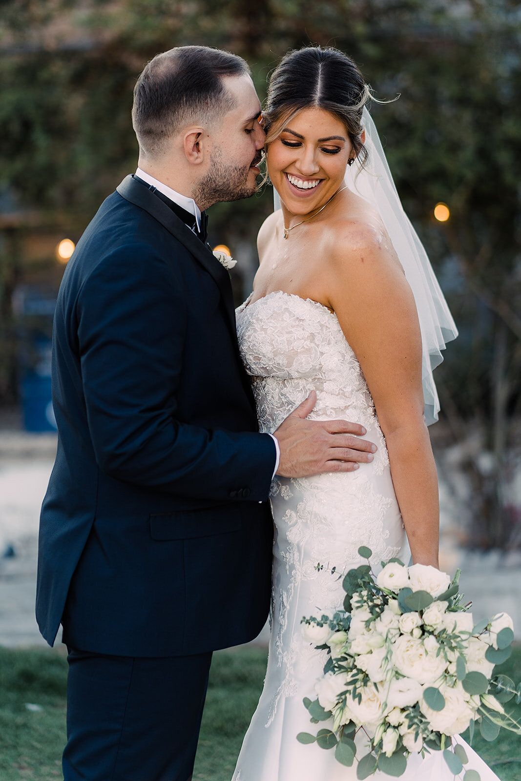A groom in a dark suit gently kisses his smiling bride on the cheek, who holds a lush bouquet of white roses