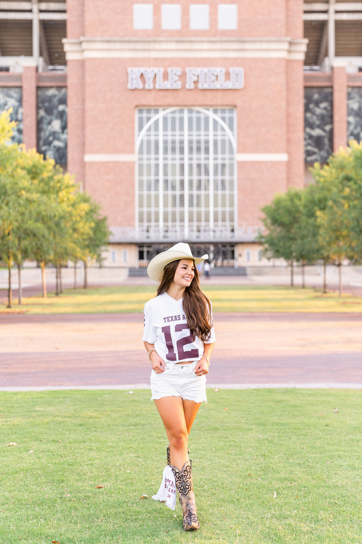 Texas A&M senior girl walking with hands in pockets and looking away while wearing cowboy hat, white Aggie jersey in front of Kyle Field at Aggie Park