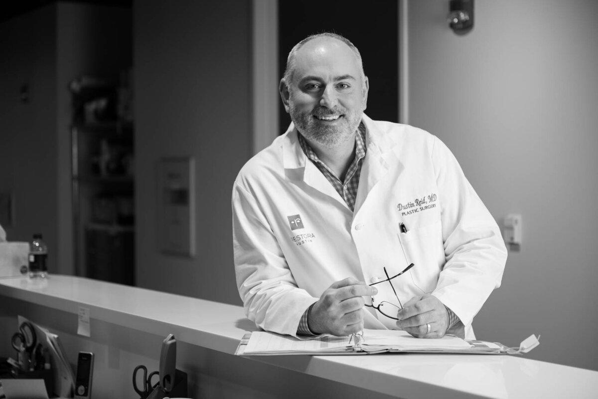 A professional image of a doctor wearing a white coat standing next to a front desk smailing to the camerra