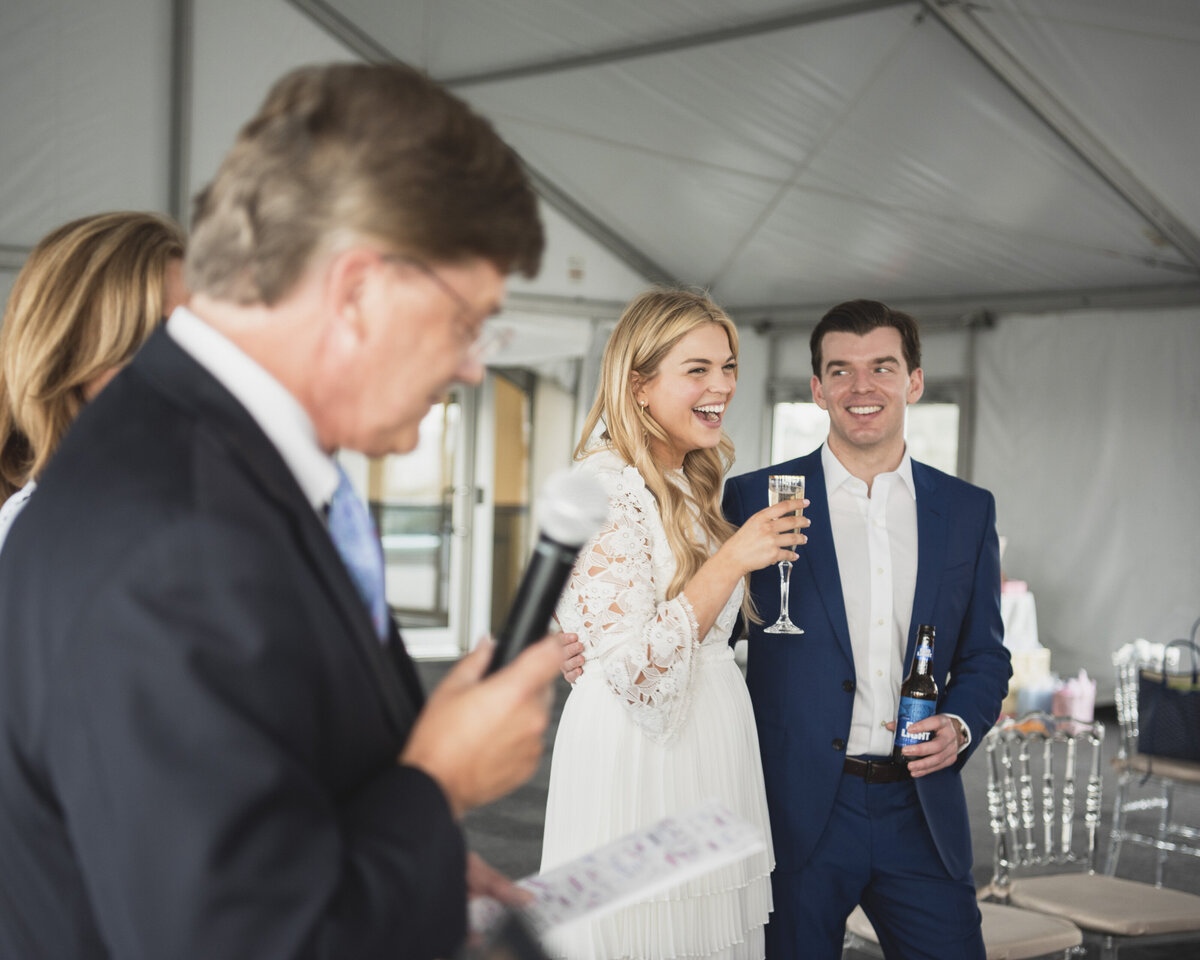 A couple holds drinks while at their engagement party. A person in the foreground holds a microphone.