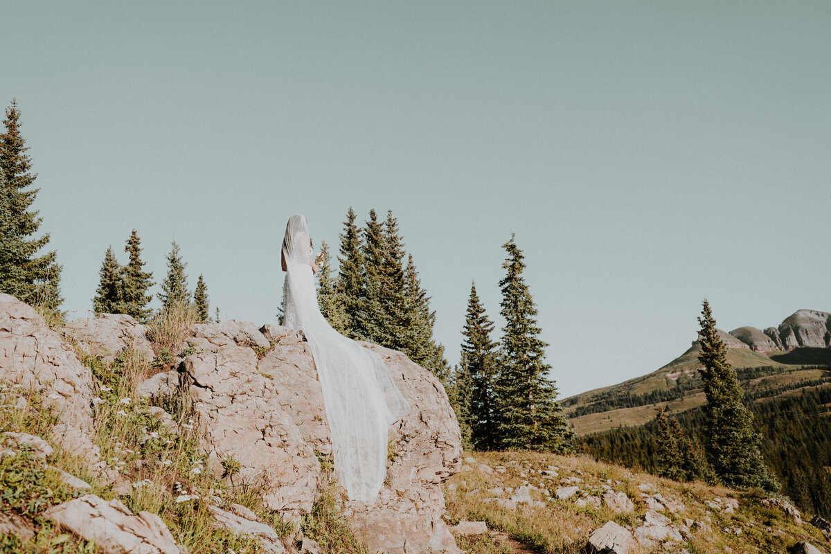 Bride with long veil stands on the edge of a rock in the mountains at sunrise before her ouray, Colorado wedding