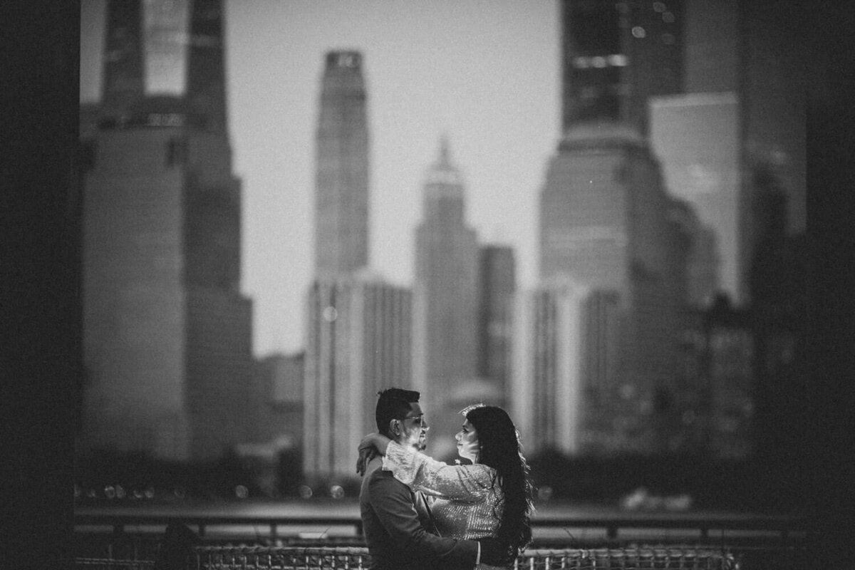 A black and white photo showing a couple hugging in New Jersey lit up by the city's skyline.