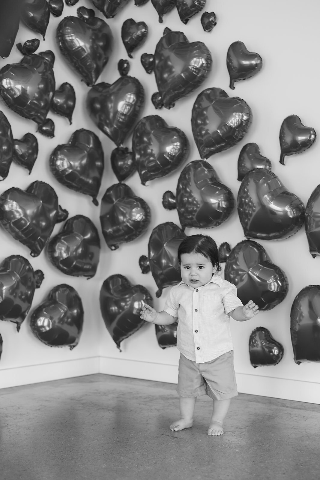 A baby boy stands in front of red heart balloons.