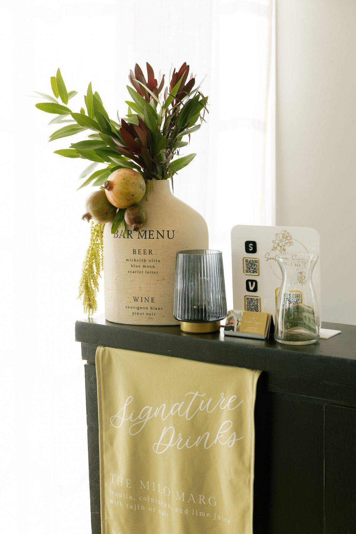 Wedding bar setup with ceramic bar menu vase filled with greenery and pomegranates next to tip jar, candles, and signature drink signage inside a bright reception space.