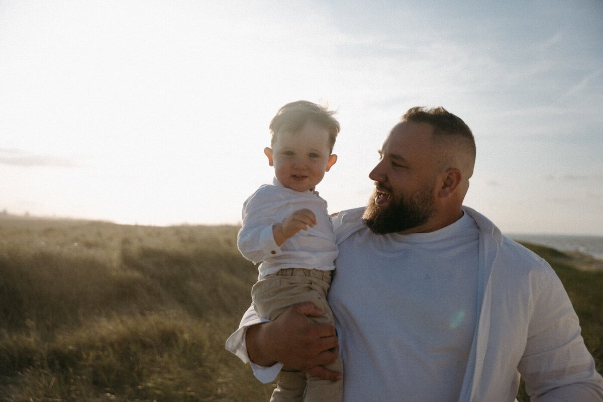 Père tenant son petit garçon dans les bras au coucher du soleil, éclats de rire complices. Reportage photo famille en bord de mer par Kamesco