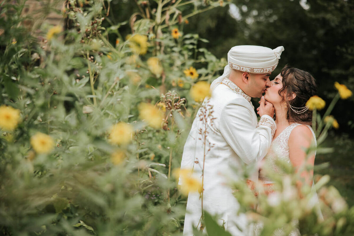 A newlywed couple shares a kiss in a lush garden, framed by vibrant yellow flowers. The groom, in a white sherwani and turban, gently holds the bride’s chin as she leans in, wearing an elegant embellished gown.