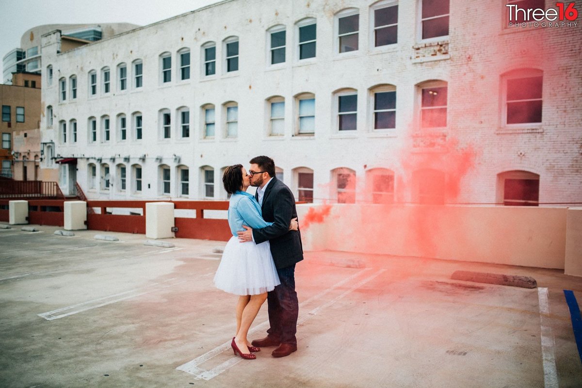 Engaged couple kiss while a red smoke illuminates the photo