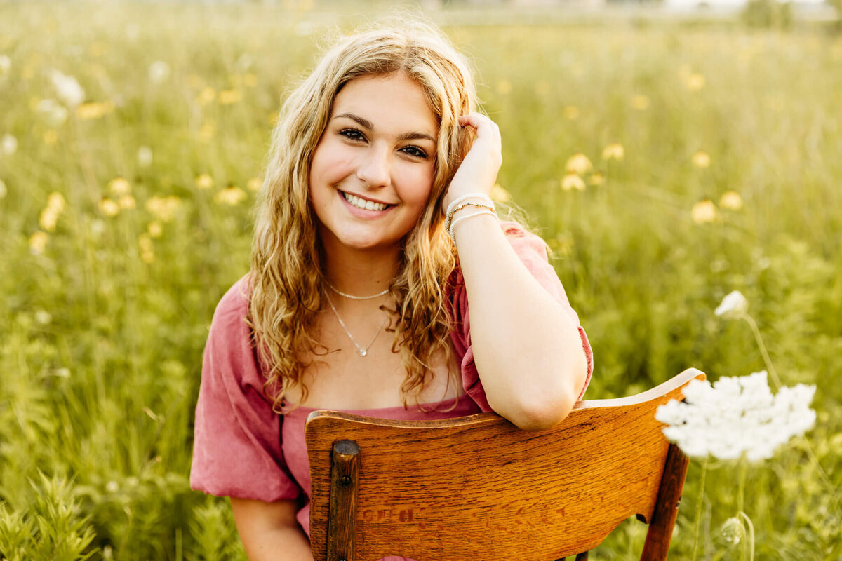 blonde high school girl leaning head against hand as arm rest on wooden chair near Oshkosh