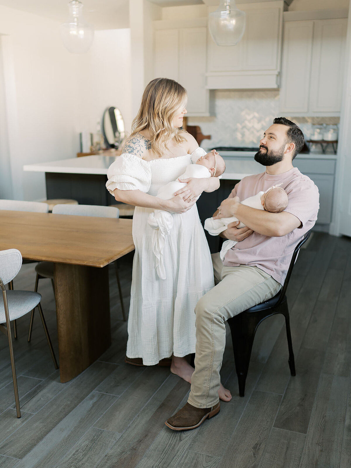 A loving couple stands together in their kitchen, each holding one of their newborn twins during a lifestyle newborn session. They look at their babies with joy and admiration, surrounded by the warmth of their home. The soft natural light highlights the intimate and tender moment of their growing family.