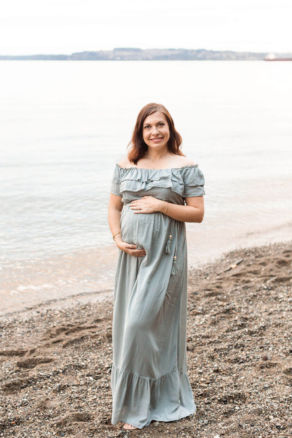 Pregnant Woman holding her belly and looking and smiling at the camera. She is wearing a long sage green dress that is off-the-shoulder.  She is standing next to a body of water.