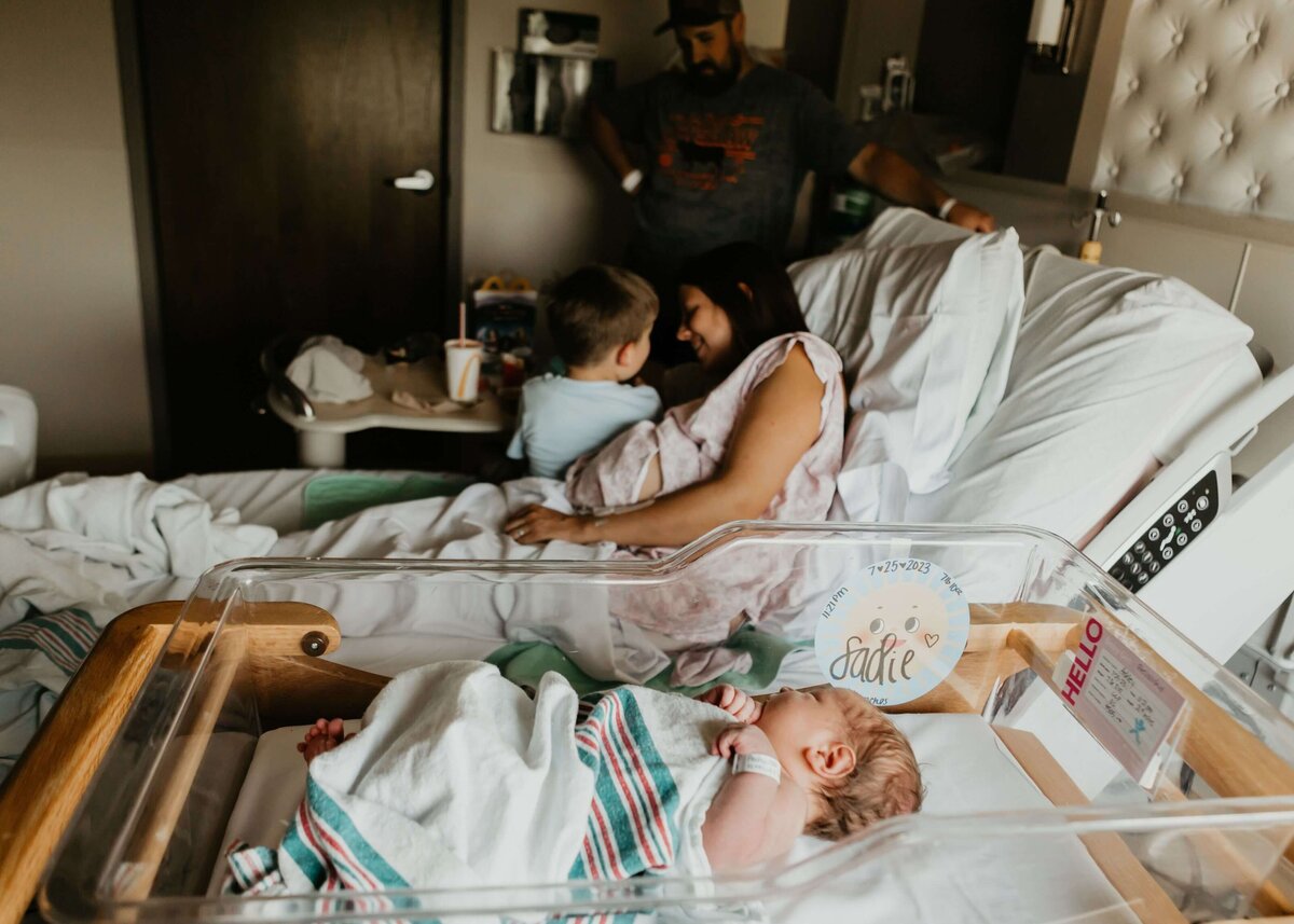 A mother holds her baby in a hospital bed while her husband and son relax in the background.