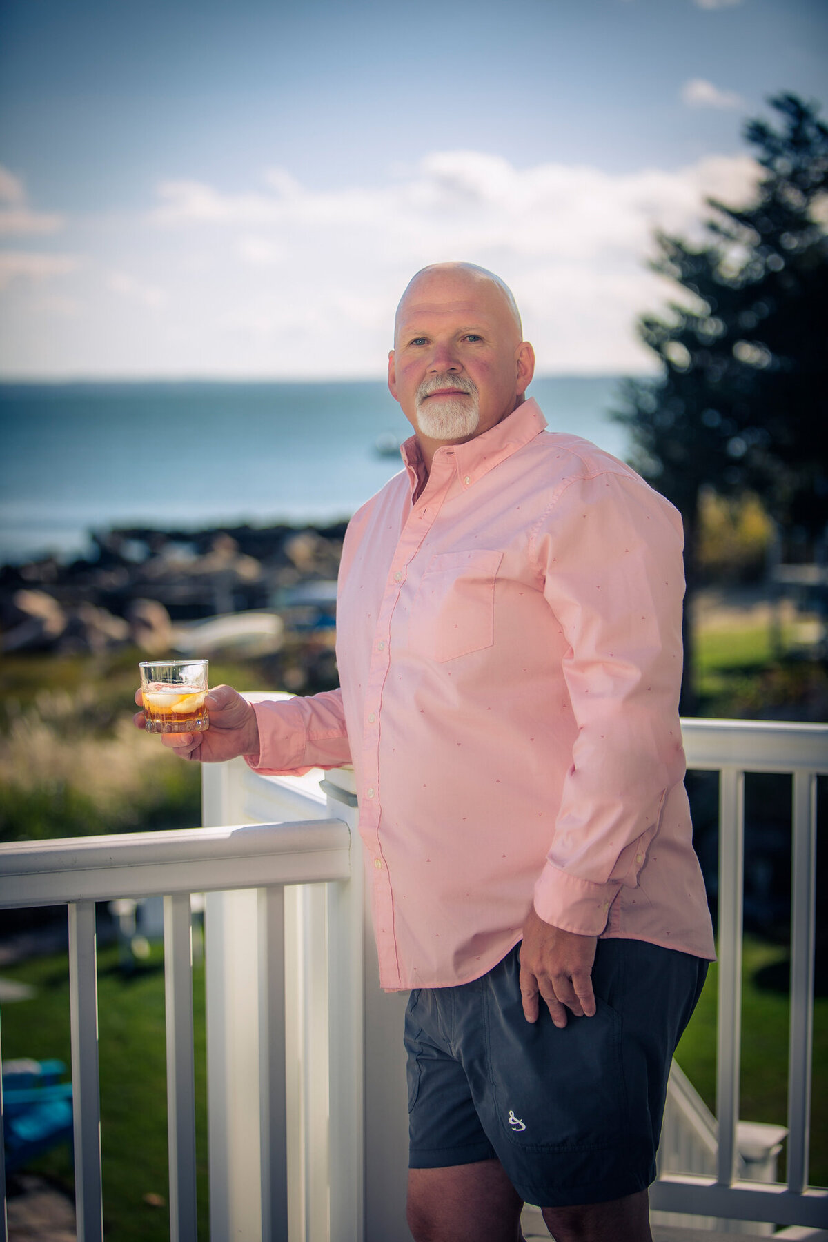 A male dating portrait client standing on his back porch in a pink dress shirt holding a whiskey