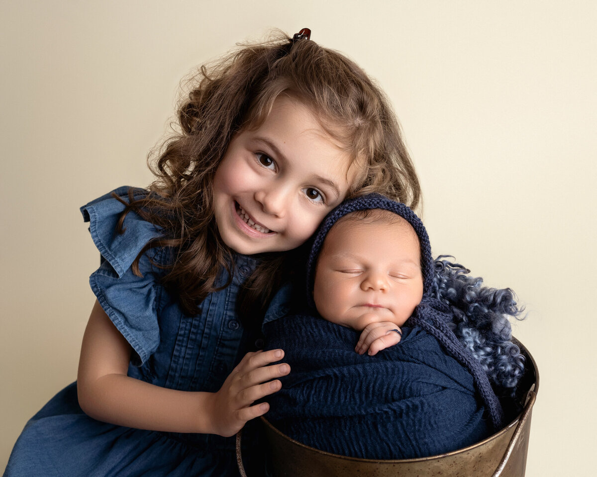 Big sister smiling and hugging her newborn brother wrapped in navy and bonnet | Sweet sibling newborn photo in Cedar Park and Austin