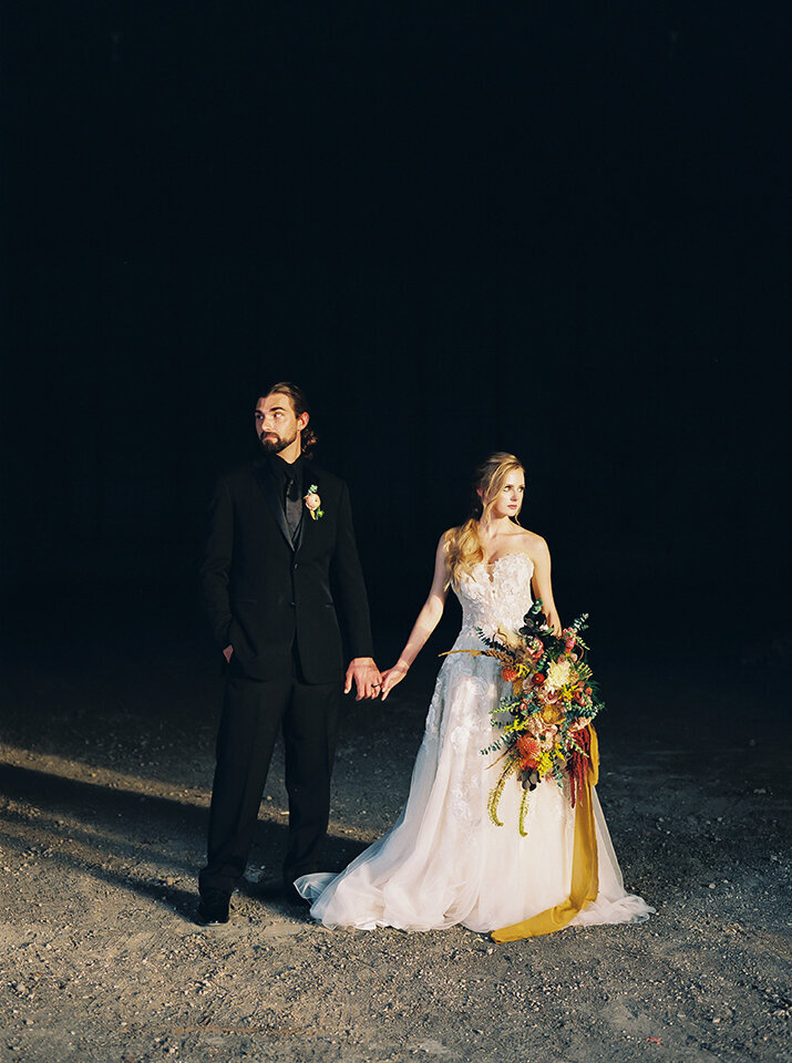 Bride and groom wearing black tuxedo and white wedding gown holding hands and a bouquet of flowers on a gravel road.