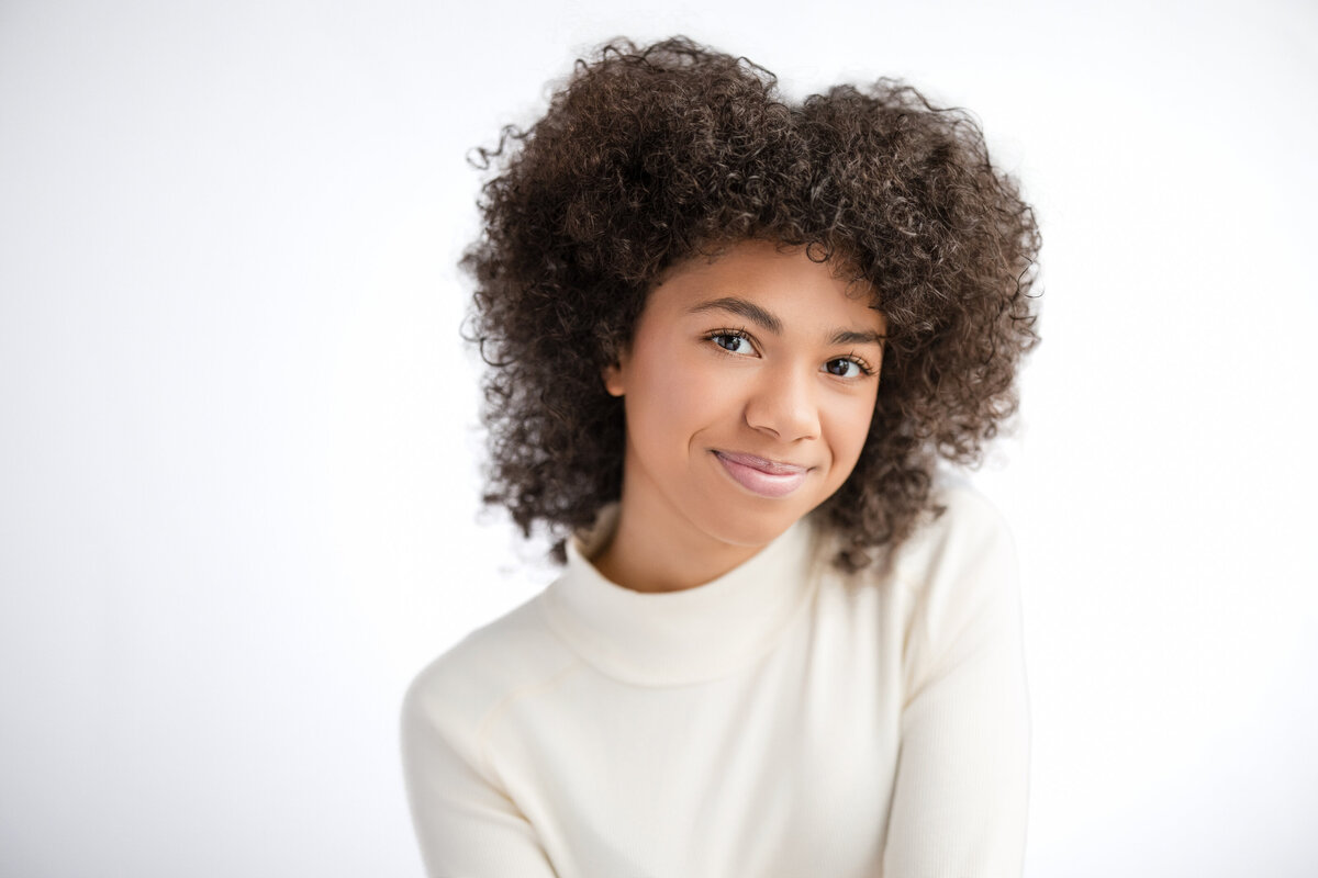 Child smiling softly in a bright studio setting, wearing a cream-colored turtleneck. Their curly hair frames their face, and they are looking directly at the camera. Clean, simple background emphasizes the subject’s expression. Kids headshots NYC.