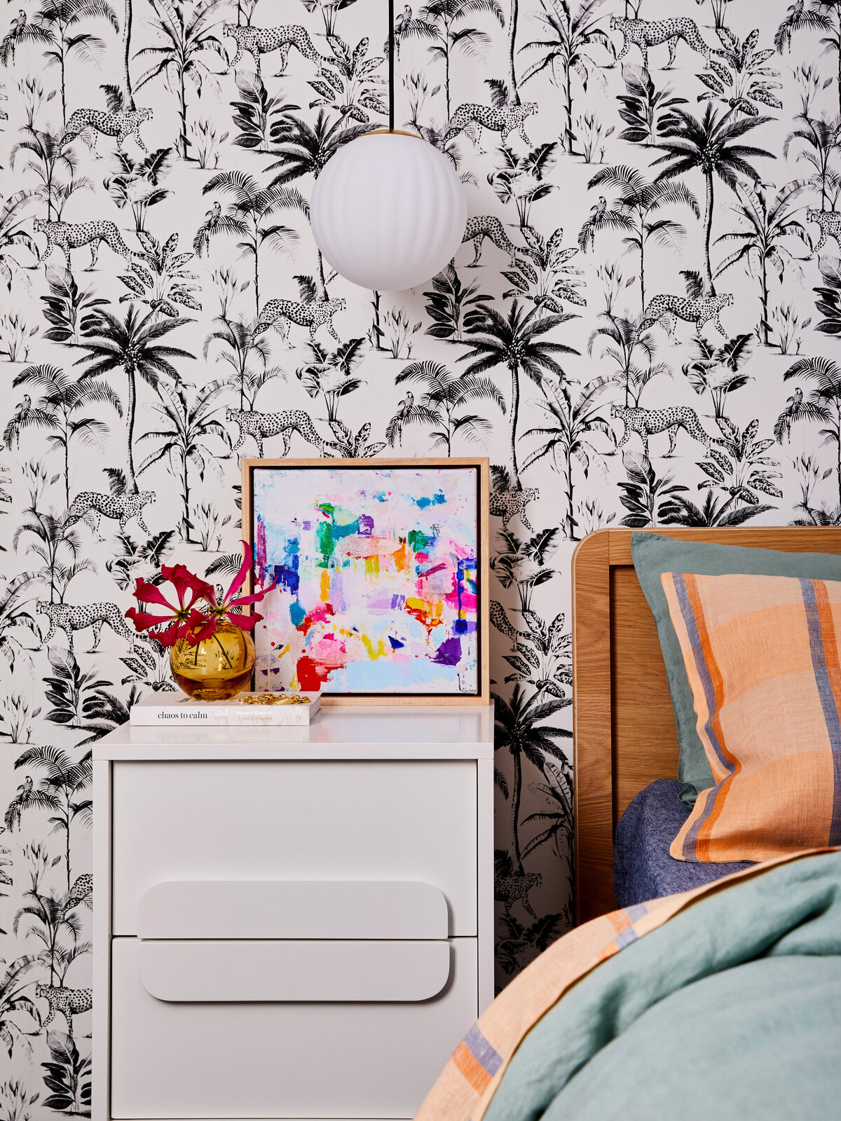A bedroom in a Sandringham home featuring bold black and white wallpaper with cheetahs and palm trees. A white bedside table is styled with a framed abstract painting and a small gold vase.