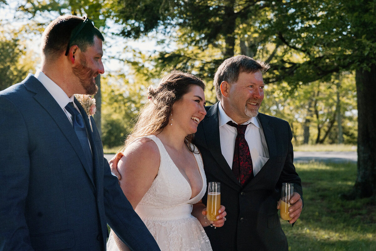 A dad has his arm around his daughter while they listen to a speech given by the mother of the bride. The groom is looking at the bride smiling. 