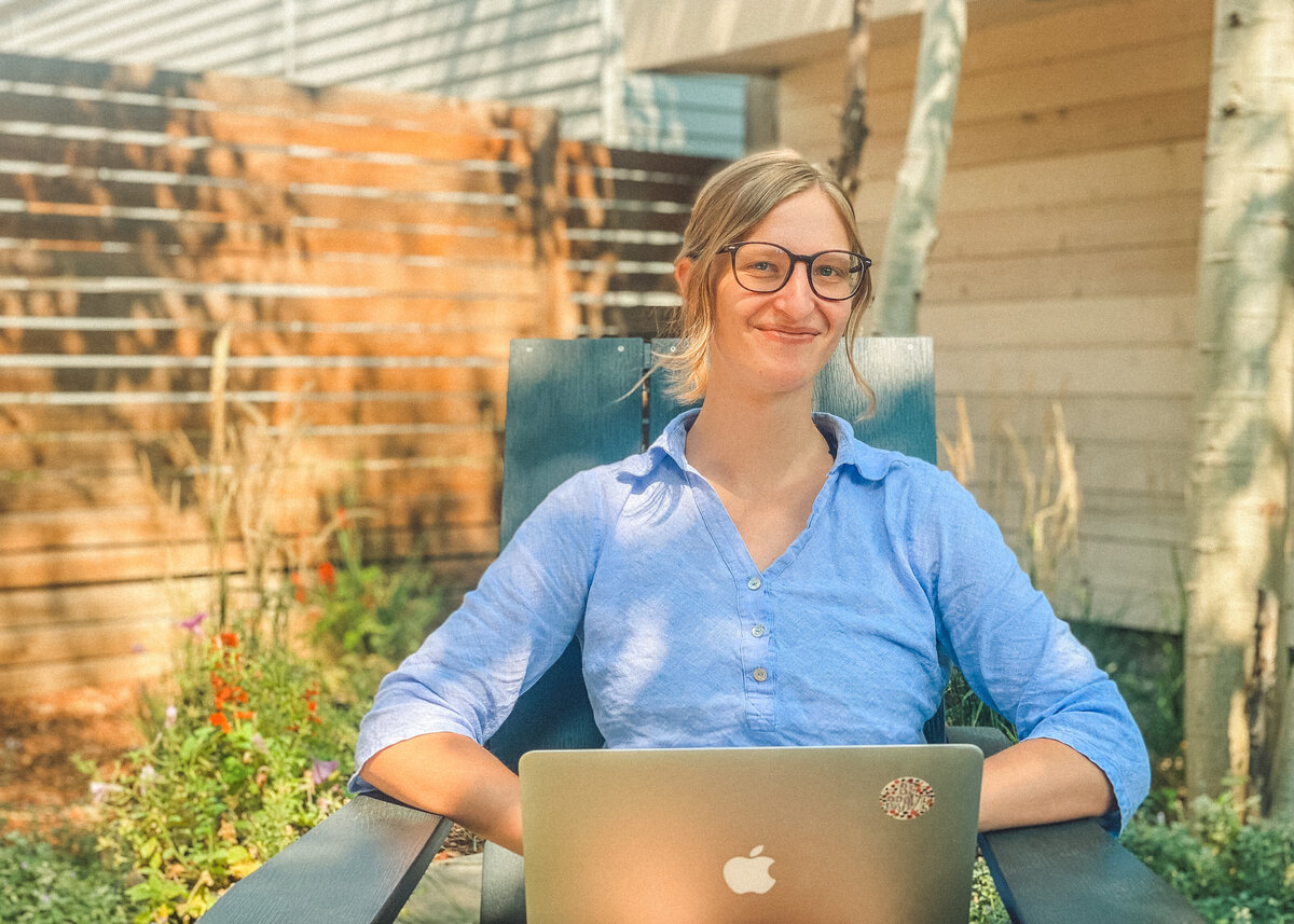 Kaitlyn Hickmann, founder of Unstoppable Copy, sits in an Adirondack chair with her open laptop and smiles. She is a white woman wearing a periwinkle collared shirt and glasses