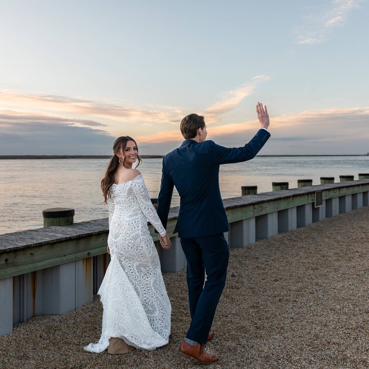 Bride and groom walk hand in hand down dock by atlantic ocean at sunset, the bride is looking over her shoulder at the camera and the groom is waving goodbye to unseen wedding guests